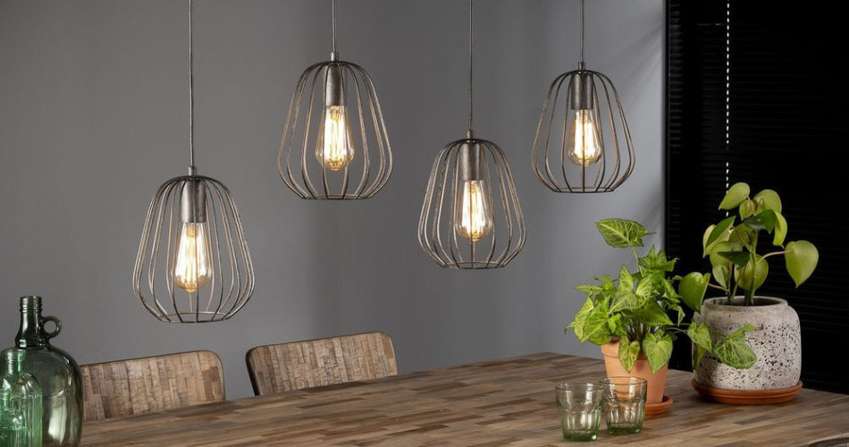Zu sehen ist eine Lampenhalterung mit 4 Lampen daran die umgeben sind von einem Vasen förmigen Gitter und diese hängen über einen hölzernen Esstisch.
